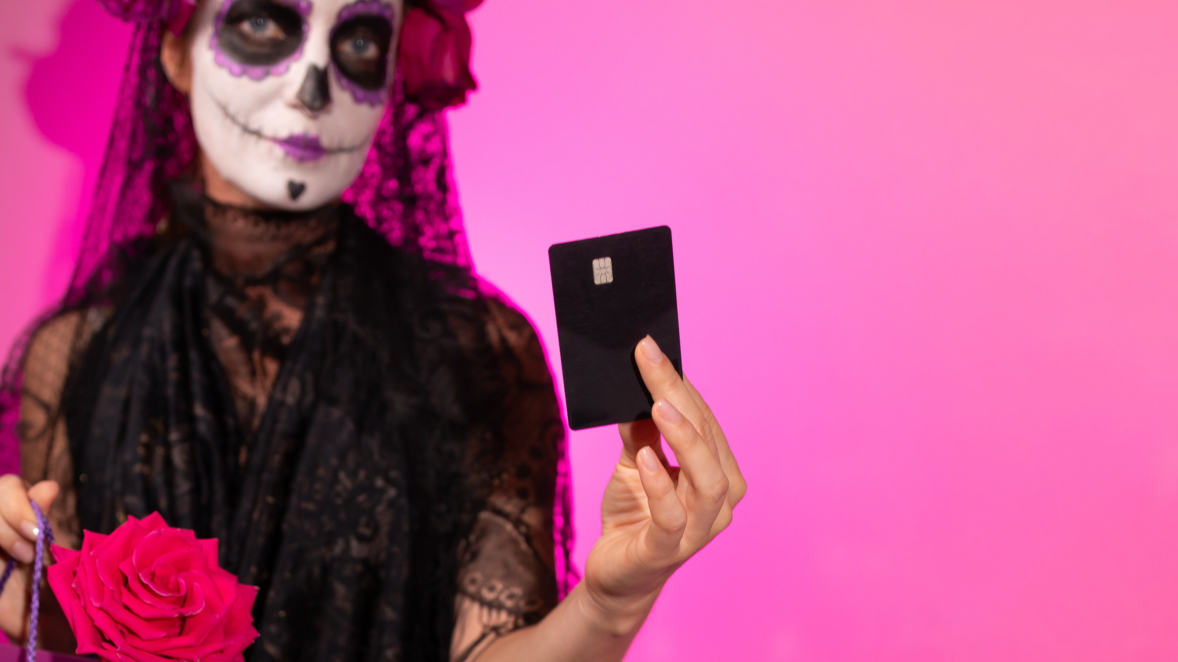 A woman dressed in a Calavera Catalina costume and wearing elaborate sugar skull makeup holds a black credit card against a bright pink background.